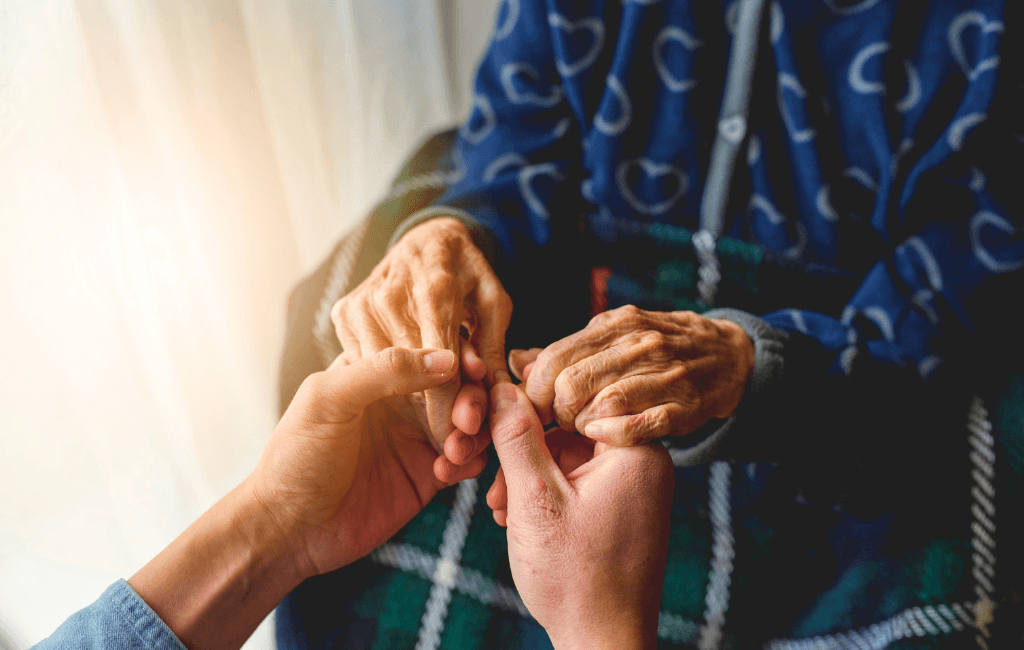 A younger person gently holding the hands of an elderly person wrapped in a plaid blanket.
