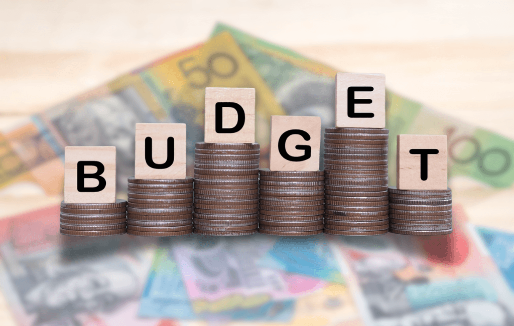 Wooden letter blocks spelling BUDGET stacked on rising piles of Australian coins with Australian currency notes in the background, representing the 2026 Federal Budget tax changes.